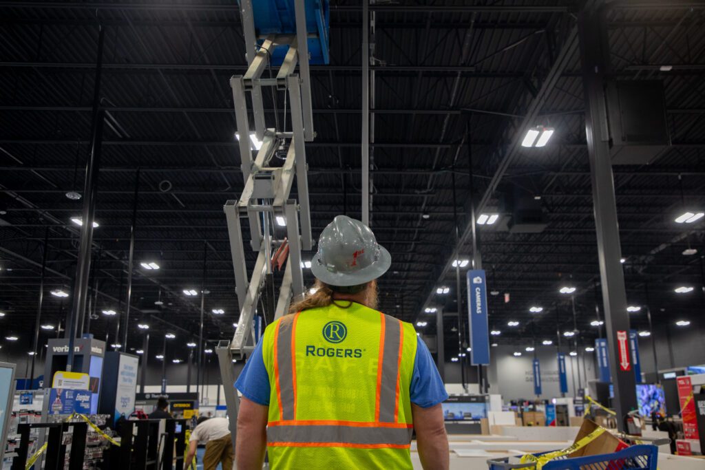 backside of an electrician with Rogers safety vest and hard hat inside a Best Buy looking at a lift to the ceiling lights