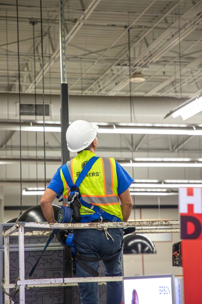 electrician in warehouse strapped to scissor lift with harness on with back facing camera with Rogers PPE gear on