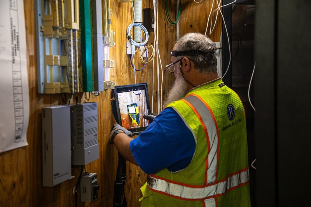 electrician taking a picture of an electrical box in a dark electrical room with PPE gear on 