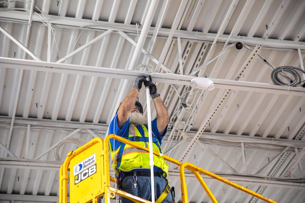 Rogers electrician inside a warehouse installing some wiring in a ceiling on a yellow scissor lift